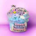 A decorative container labeled 'Candy Kitten Clouds' filled with pastel blue and lavender fluffy material, topped with sprinkles, candy-like beads, a small cat figurine, and a tag that reads 'Scoop'. The product is photographed against a pink-to-purple gradient studio background.