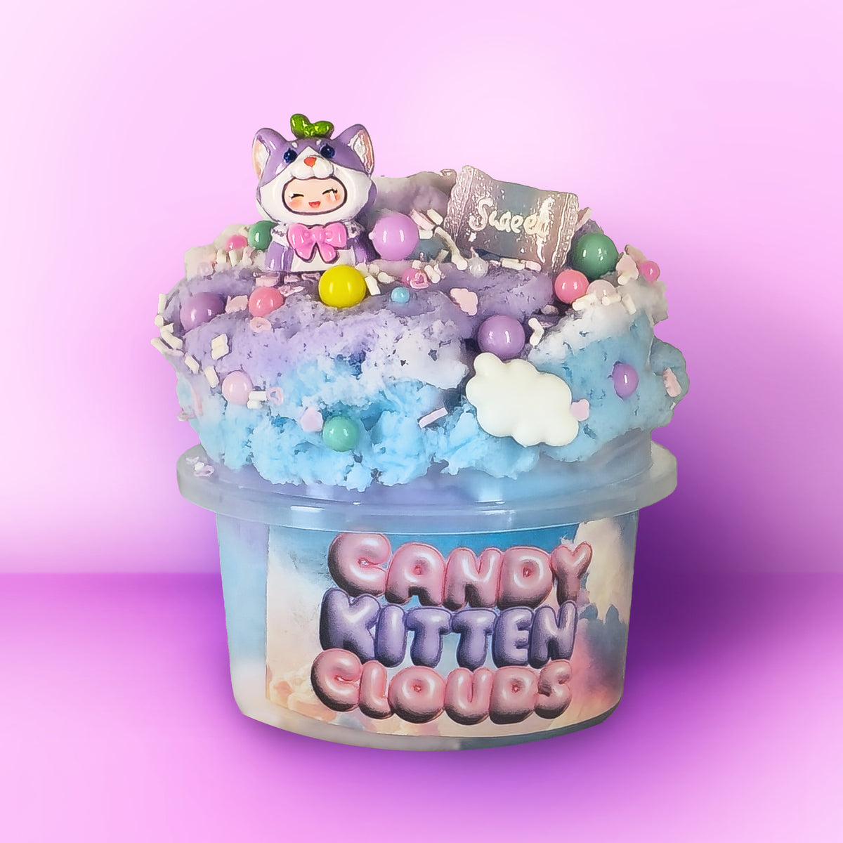 A decorative container labeled 'Candy Kitten Clouds' filled with pastel blue and lavender fluffy material, topped with sprinkles, candy-like beads, a small cat figurine, and a tag that reads 'Scoop'. The product is photographed against a pink-to-purple gradient studio background.