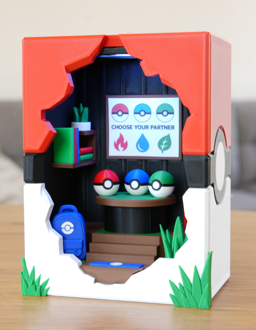 A 3D-printed Pokémon-themed miniature display box or room, featuring a red and white Pokémon Ball design with a cutout front showing a detailed interior with a screen, Pokémon Balls, a blue backpack, and green grass accents.