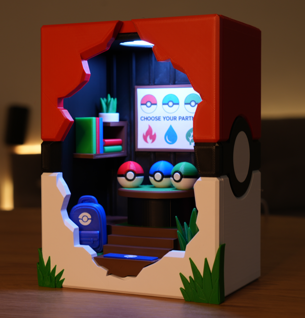 A miniature diorama set inside a Pokéball-shaped box, featuring a small room with shelves, a screen reading 'CHOOSE YOUR PARTY', three colored Pokéball eggs, a blue Pokéball backpack, and green grass accents, photographed on a tabletop in a dim indoor setting.