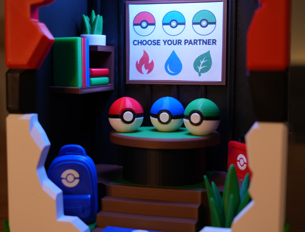 A miniature diorama setup featuring three Pokéball-style eggs on a table with a sign reading 'CHOOSE YOUR PARTNER' above them, alongside a blue Pokéball backpack and themed shelving in a dark room.