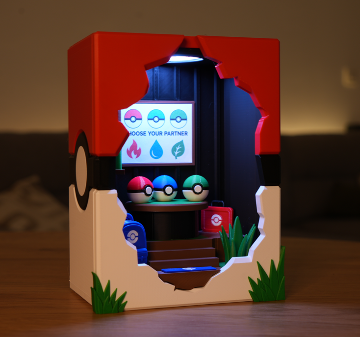 A Pokémon-themed miniature diorama set inside a Pokéball-shaped box, featuring a red and black top, beige lower half, a lit screen, three Pokéballs, a small red bag, stairs, and green grass accents on a wooden surface.