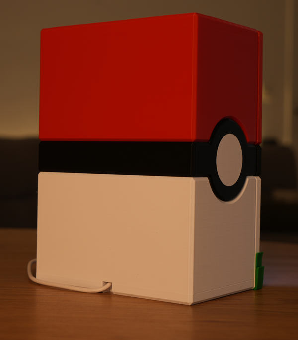 A 3D-printed toy box designed to look like a classic Pokéball, featuring a red top half, a black horizontal band with a white center circle, and a white bottom half, sitting on a wooden surface.