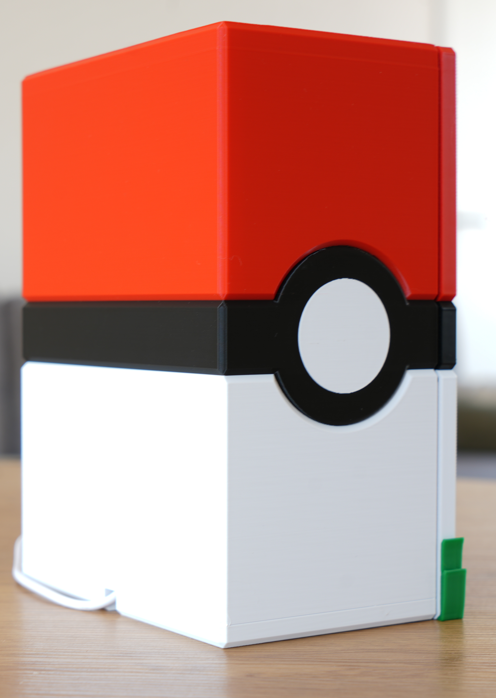 A 3D-printed toy box designed to look like a Pokéball, featuring a red top half, a black and white center band with a circular emblem, and a white bottom half, shown sitting on a wooden table.