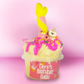 A novelty toy or container labeled 'Derp's Birthday Bash' filled with pale yellow material and decorated with bright pink icing-like drizzle, sprinkles, a yellow mermaid tail, and a small white cat figurine, set against a pink gradient background.
