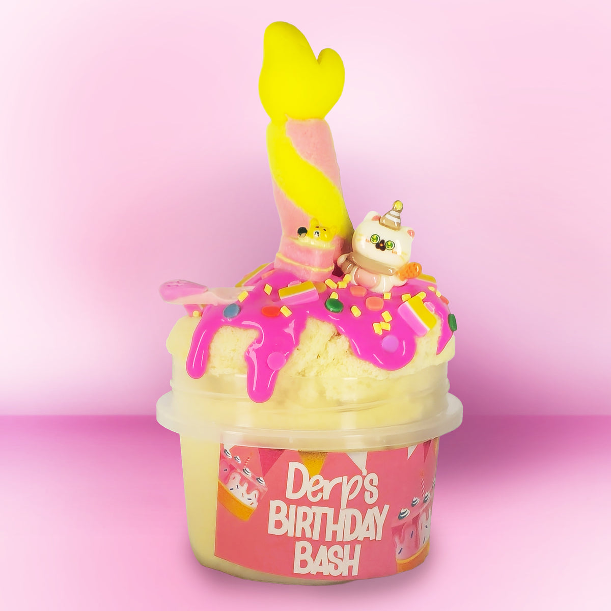 A novelty toy or container labeled 'Derp's Birthday Bash' filled with pale yellow material and decorated with bright pink icing-like drizzle, sprinkles, a yellow mermaid tail, and a small white cat figurine, set against a pink gradient background.