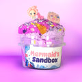 A clear plastic tub labeled 'Mermaid's Sandbox' filled with bright pink and purple sand, topped with small mermaid figurines and colorful decorative pieces, set against a pink gradient background.