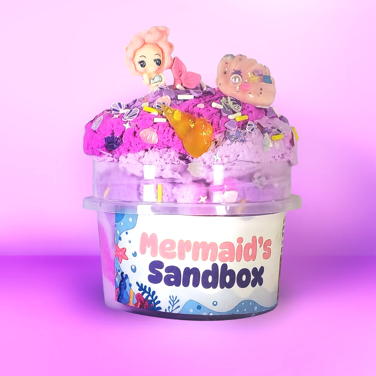 A clear plastic tub labeled 'Mermaid's Sandbox' filled with bright pink and purple sand, topped with small mermaid figurines and colorful decorative pieces, set against a pink gradient background.