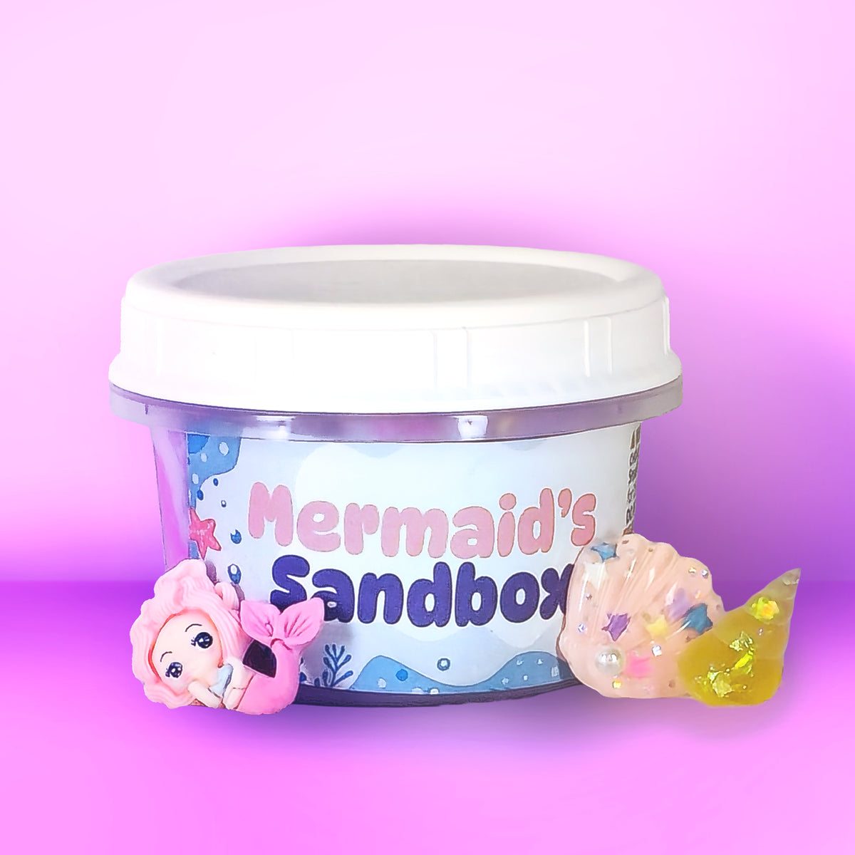 A clear plastic tub with a white lid labeled 'Mermaid's Sandbox' on a pink gradient background, with a pink mermaid figure and a translucent yellow seashell toy placed in front.