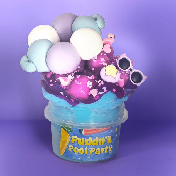 A colorful slime bucket toy from Puddn's Pool Party, featuring a blue base, purple and pink slime, and various decorative clay-like accessories including pastel balls, sunglasses, and a small pink flamingo, set against a solid purple background.