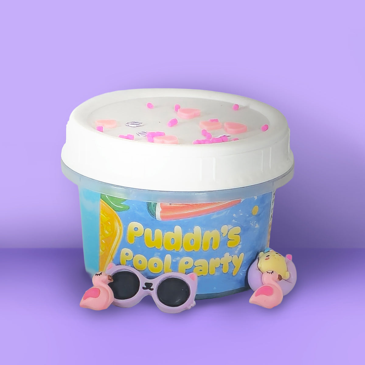 A clear plastic tub with a white lid, filled with a blue gel or slime-like product and decorated with pink heart sprinkles. The tub has a colorful label reading 'Puddn's Pool Party' with a flamingo and pool graphic.