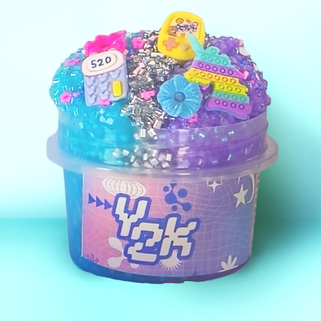 A small clear plastic tub filled with decorative slime or craft material, topped with colorful faux bricks, beads, glitter, and small toy-like embellishments. The tub has a pink-to-purple label with large stylized text reading Y2K.