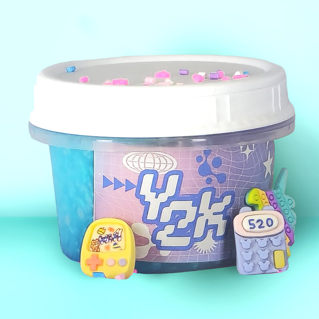 A clear plastic tub of decorative slime with a white lid, featuring a blue and purple printed label with the text 'YOK' and small pastel decorative pieces on top. Two small toy-like accessories are placed in front of the tub on a light aqua background.